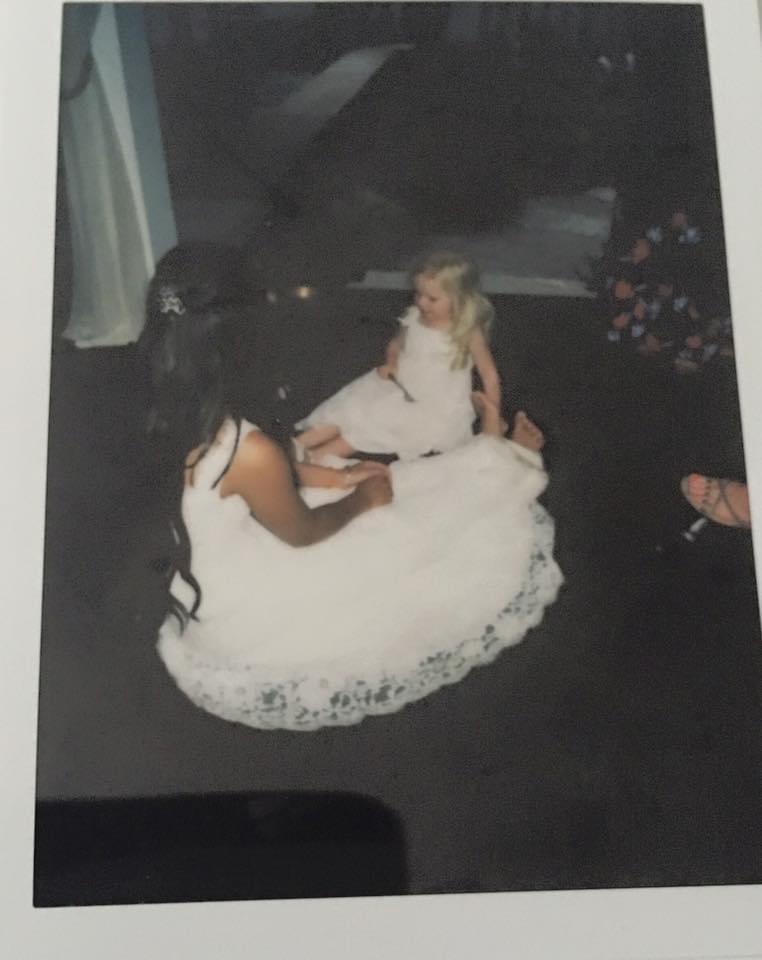 Nina in her wedding dress sitting on the ground with Abigail in her flower girls dress. Nina sent this picture.